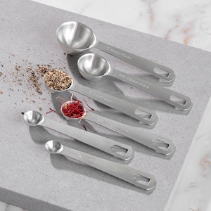 May include: Set of five stainless steel measuring spoons with different sizes. The spoons are arranged in a row on a gray surface. The spoons are labeled with measurements in both teaspoons and milliliters. The spoons are filled with various spices, including black peppercorns, saffron threads, and a blend of herbs.