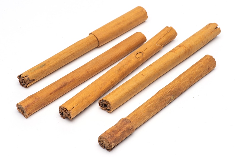 May include: Five cinnamon sticks of varying lengths, arranged in a fan shape on a white background.