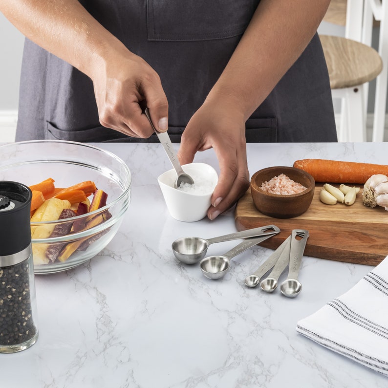 May include: A set of six stainless steel measuring spoons on a white countertop. The spoons are arranged in a row, with the largest spoon on the left and the smallest spoon on the right. The spoons are all shiny and new.