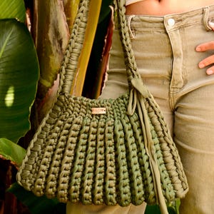 May include: Olive green crochet handbag with braided handles and a rectangular body. The bag features a wooden tag with initials. The bag is made of a thick, textured yarn and has a soft, draped appearance.