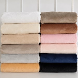 May include: A stack of twelve folded plush blankets in various colors labeled White, Ivory, Oatmeal, Taupe, Silver, Honey, Mocha, Chestnut, Baby Pink, Peach n Cream, Navy, and Black. Each blanket has a soft, uniform texture and is neatly stacked.