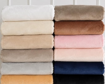 May include: A stack of twelve folded plush blankets in various colors labeled White, Ivory, Oatmeal, Taupe, Silver, Honey, Mocha, Chestnut, Baby Pink, Peach n Cream, Navy, and Black. Each blanket has a soft, uniform texture and is neatly stacked.