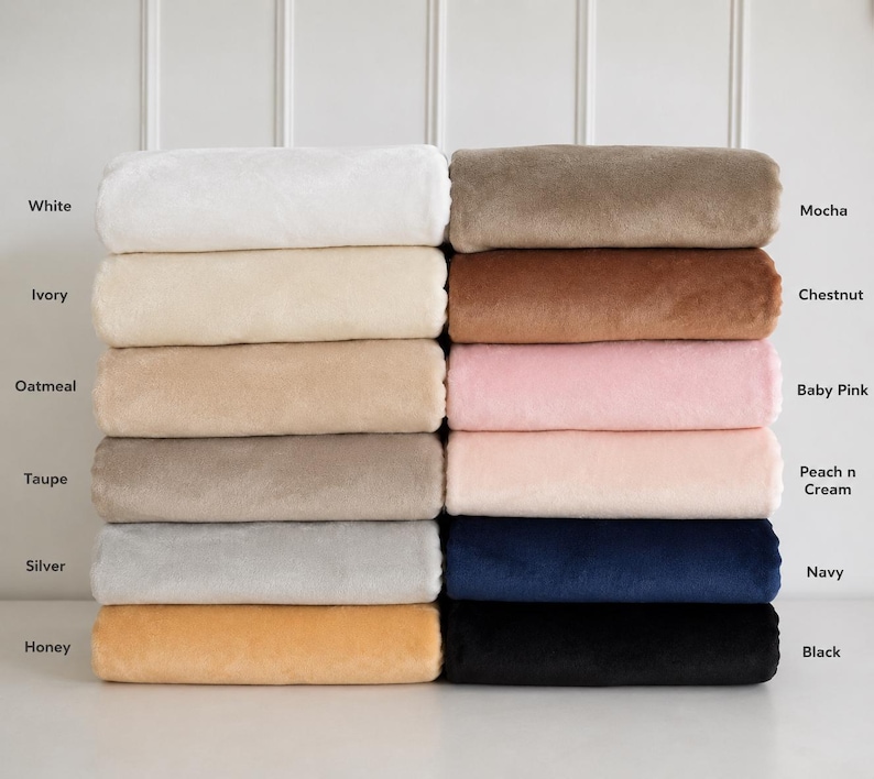 May include: A stack of twelve folded plush blankets in various colors labeled White, Ivory, Oatmeal, Taupe, Silver, Honey, Mocha, Chestnut, Baby Pink, Peach n Cream, Navy, and Black. Each blanket has a soft, uniform texture and is neatly stacked.