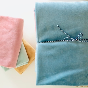 May include: A stack of folded velvet fabric pieces in various colors including dusty pink, light mint green, mustard yellow, and a large light blue piece tied with a blue and white baker's twine string. The fabric has a soft, plush texture.