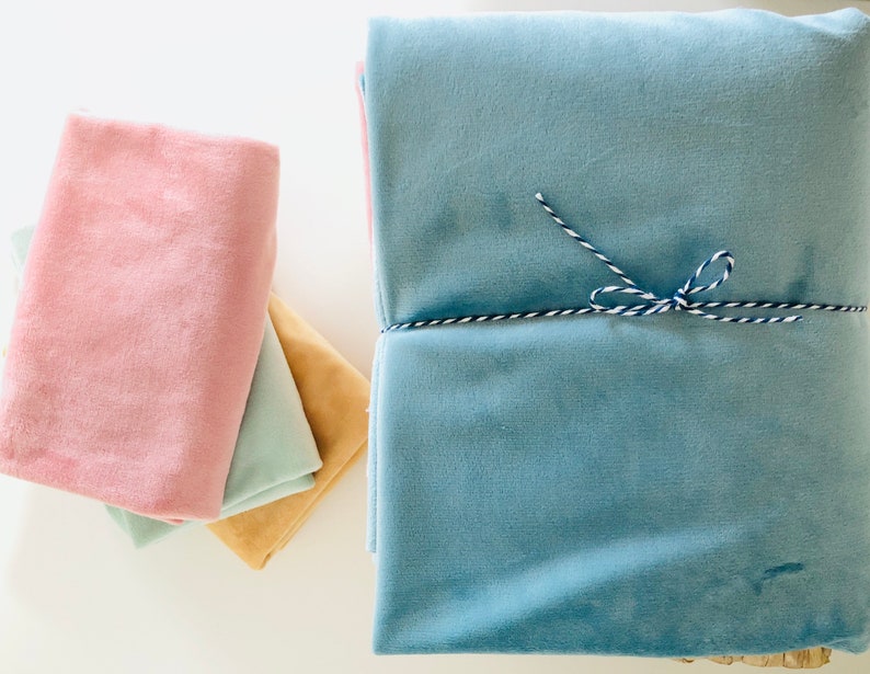 May include: A stack of folded velvet fabric pieces in various colors including dusty pink, light mint green, mustard yellow, and a large light blue piece tied with a blue and white baker's twine string. The fabric has a soft, plush texture.