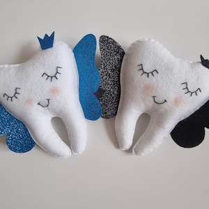 May include: Two plush white teeth with embroidered eyes and smiles, each with a pair of wings. One tooth has blue glitter wings and a blue crown, the other has black glitter wings and a black bow.