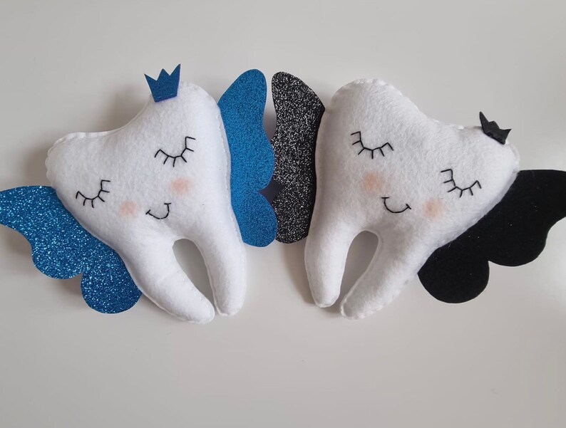 May include: Two plush white teeth with embroidered eyes and smiles, each with a pair of wings. One tooth has blue glitter wings and a blue crown, the other has black glitter wings and a black bow.