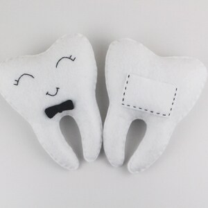 May include: Two white felt tooth pillows. One tooth has a black bow tie and a smiling face. The other tooth has a blank white rectangle with black stitching.
