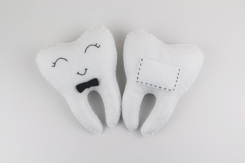 May include: Two white felt tooth pillows. One tooth has a black bow tie and a smiling face. The other tooth has a blank white rectangle with black stitching.