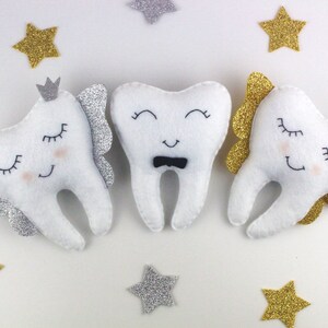 May include: Three white felt tooth-shaped plush toys with embroidered eyes and mouths. The middle tooth has a black bow tie. The tooth on the left has silver glitter wings and a crown. The tooth on the right has gold glitter wings and a crown.