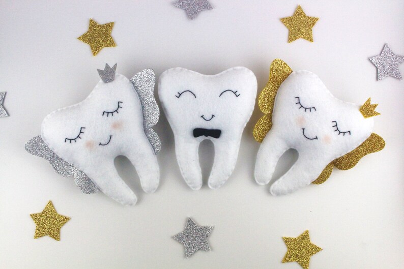 May include: Three white felt tooth-shaped plush toys with embroidered eyes and mouths. The middle tooth has a black bow tie. The tooth on the left has silver glitter wings and a crown. The tooth on the right has gold glitter wings and a crown.