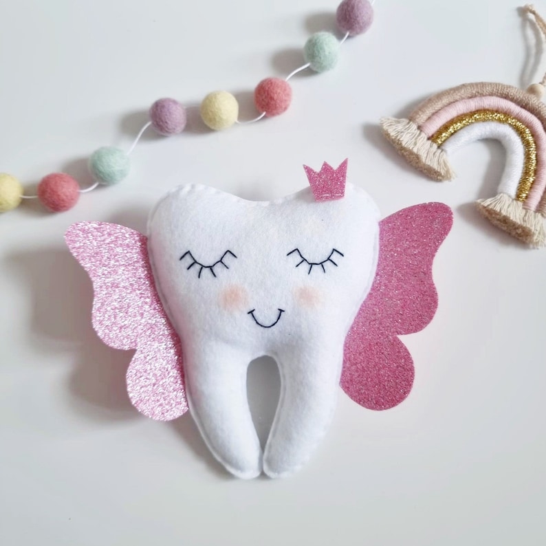 May include: A white felt tooth with pink glitter wings and a small pink crown on top. The tooth has a smiling face with closed eyes.