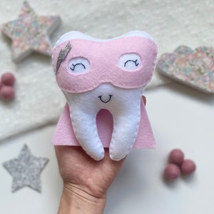 May include: A white felt tooth with a pink superhero mask and cape. The tooth has a smiling face and a silver lightning bolt detail on the mask. Decorative stars and felt balls are in the background.