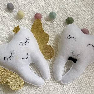 May include: Two white felt tooth-shaped plush toys with embroidered eyes and smiles. One tooth has a gold glitter crown and wings, the other has a black bow tie.