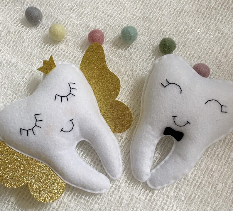 May include: Two white felt tooth-shaped plush toys with embroidered eyes and smiles. One tooth has a gold glitter crown and wings, the other has a black bow tie.
