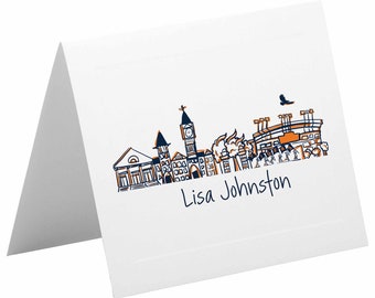 May include: A white folded card with a hand-drawn illustration of a city skyline in orange and blue. The skyline includes a stadium, a church, and several buildings. The text "Lisa Johnston" is written in blue ink below the illustration.