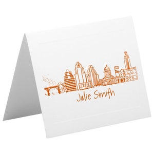 Puede incluir: Una tarjeta blanca doblada con un dibujo de línea naranja del horizonte de Austin, Texas. El texto "Julie Smith" está impreso debajo del horizonte.