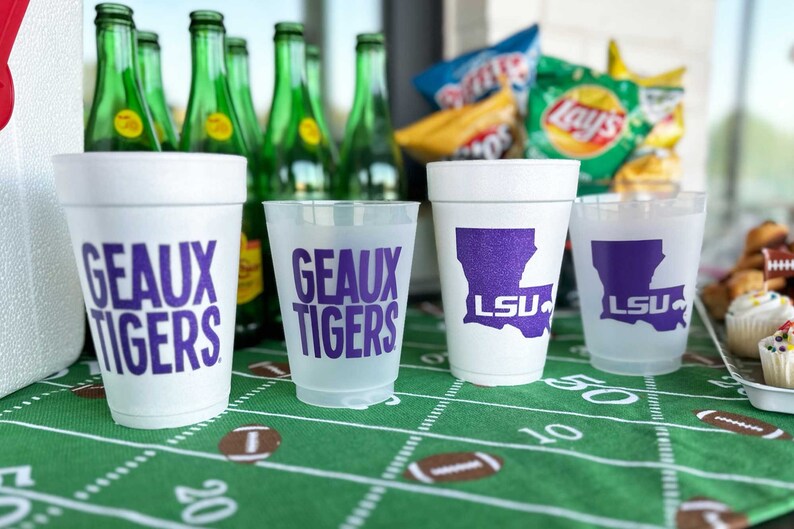 May include: White foam cups and clear plastic cups with purple text and logos. The cups feature the words "GEAUX TIGERS" and the LSU logo. Green glass bottles and snacks are in the background, on a green football field-themed tablecloth.