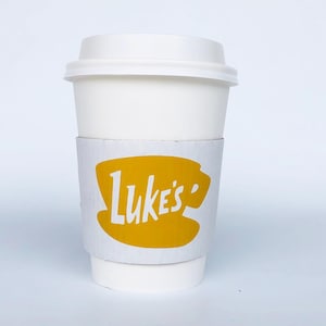 以下が含まれることがあります： 白い蓋付きの紙コップ。黄色いスリーブには白い文字で「Luke's」と書かれています。ホットドリンク用のカップです。