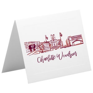 Puede incluir: Una tarjeta blanca doblada con un dibujo a tinta marrón de una ciudad. El dibujo incluye un estadio, edificios y un puente. El texto "Charlotte Woodson" está escrito en tinta marrón debajo del dibujo.