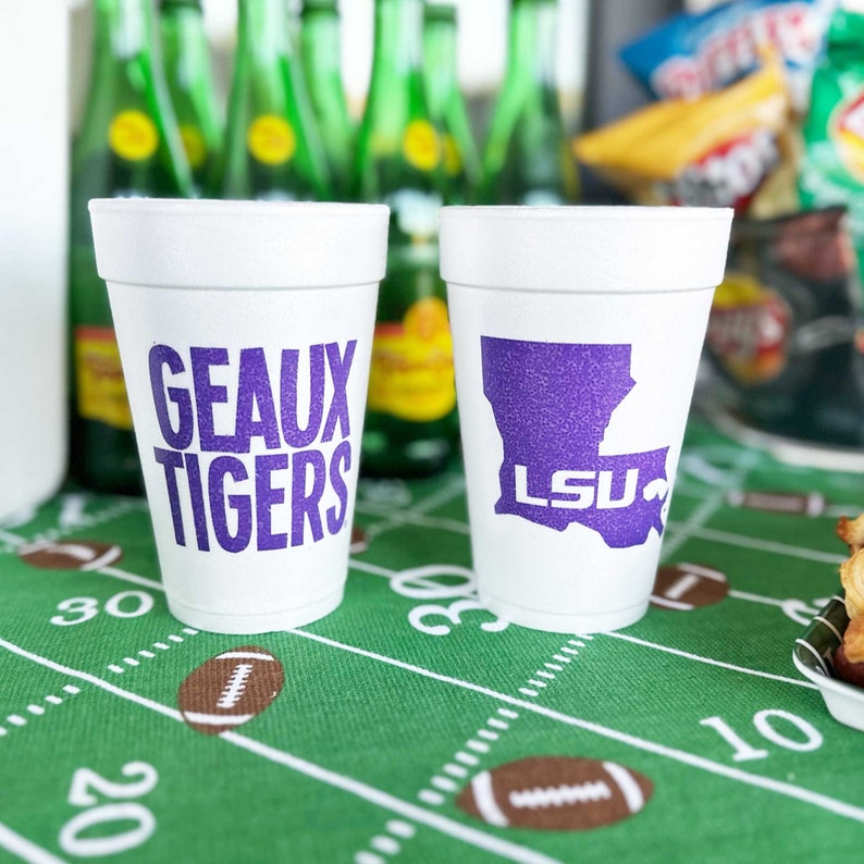 May include: Two white foam cups with purple text and graphics. One cup reads "GEAUX TIGERS", the other features a purple outline of Louisiana with "LSU" inside. The cups sit on a green football field-themed tablecloth.