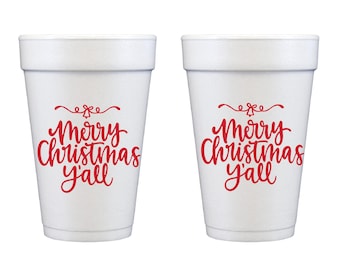 Puede incluir: Dos vasos desechables de espuma blanca con texto rojo que dice Merry Christmas y'all en una fuente cursiva. Sobre el texto hay un pequeño dibujo lineal rojo de una campana con remolinos decorativos a cada lado del icono.