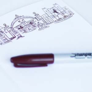 Peut inclure: Un bloc-notes blanc avec un dessin à l'encre noire d'une ville. Un marqueur permanent Sharpie brun est posé sur le bloc-notes.