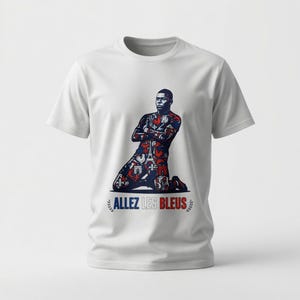 May include: White t-shirt featuring a graphic of a soccer player kneeling with arms crossed. The player's uniform is patterned with red, white, and blue designs. The text "ALLEZ LES BLEUS" is printed below the image.