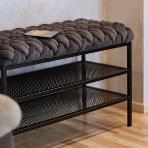 May include: A black metal bench with two shelves and a gray woven cushion top. The bench is in front of a white wall.