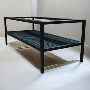 May include: A black metal coffee table with a rectangular frame and a lower shelf. The shelf has a perforated design. The table has four legs and is set against a white background.