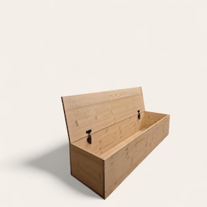 May include: A light-colored wooden storage chest with the lid partially open. The chest is rectangular with a hinged lid and metal hardware. The wood grain is visible, and the chest casts a shadow.