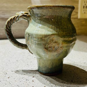 May include: Handmade ceramic mug with a textured handle and a unique, glazed finish. The mug features a light green and brown color palette, with a detailed, sculpted handle. The mug's design is both rustic and elegant, perfect for enjoying your favorite beverage.