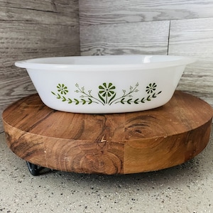 May include: White oval baking dish with a green floral design. The dish is sitting on a wooden stand.