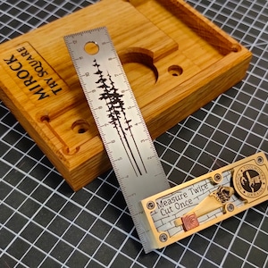 Könnte beinhalten: Ein Messing-Winkelmesser mit der Gravur "Measure Twice Cut Once" auf der Oberfläche. Das Werkzeug wird von Mirock Toolworks in der Ukraine hergestellt. Das Werkzeug liegt auf einer schwarz-weißen Gitteroberfläche neben einer Holzkiste mit einem Lineal und der Gravur "Mirock Try Square" auf der Kiste.