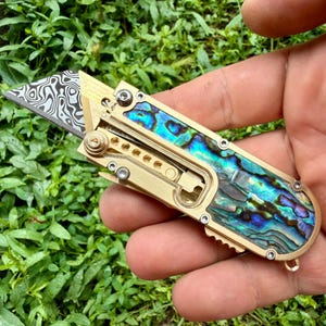 May include: A folding utility knife featuring a brass handle with iridescent abalone shell inlays. The blade displays a distinct black and silver Damascus steel pattern. The tool includes exposed mechanical screws and a small metal loop at the base.