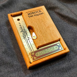 May include: A wooden try square with a brass ruler and a brass plate with the text "MIROCK" engraved on it. The try square is made of light brown wood and has a dark brown wood inlay. The ruler is marked in inches.