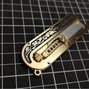 以下が含まれることがあります： 真鍮製の折りたたみ式ユーティリティナイフ。U字型のフレームには黒いインレイの上に複雑な花の彫刻が施されています。メカニズムには、円形の切り抜きがある中央の真鍮製バー、小さな調整ノブ、部分的に伸びた金属製の刃が含まれています。