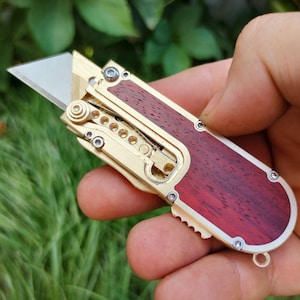 Puede incluir: Una pequeña navaja plegable con un marco de metal dorado y un mango de madera rojo oscuro. La hoja plateada está expuesta. El cuchillo se sostiene en una mano sobre un fondo verde borroso.