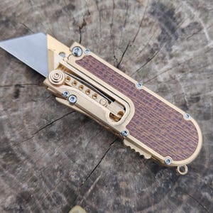 May include: A utility knife with a silver blade and a gold-coloured metal frame. The handle features a brown wood grain pattern. The knife is open, revealing the blade. The knife is on a wooden surface.