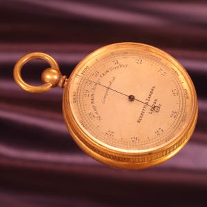 May include: A round gold-toned pocket barometer with a circular ring at the top. The face features the text Negretti & Zambra London, Compensated, and weather indicators including Stormy, Rain, Change, Fair, and Very Dry with numerical scales.