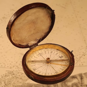 May include: A vintage compass in a brown leather case, open to show the compass face. The compass features a gold bezel with directional markings and a needle. The case is lined with a cream-coloured fabric.