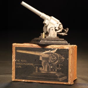 Può includere: Un cannone in miniatura color argento con una base nera è posto sopra una scatola di cartone. La scatola presenta un disegno del cannone e il testo "THE N.D.C. BREECHLOADING GUN."
