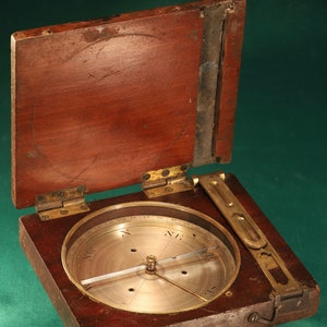 May include: A vintage wooden compass with a brass face and a hinged lid. The compass is open and the needle is pointing north.