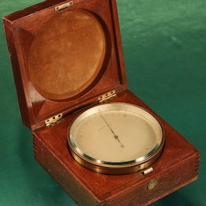 May include: A wooden box with a hinged lid, containing a brass and glass barometer with a white face and black needle. The barometer reads "Compensated".