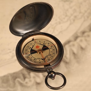 May include: An open, antique compass with a black metal casing and a hinged lid. The compass face features directional markings, a needle, and a red accent. A small ring is attached to the bottom. The background is a map.