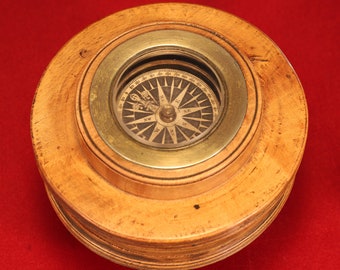 May include: A wooden compass with a brass rim and a glass face. The compass has a black needle and a white background with black markings. The compass is set in a wooden base.