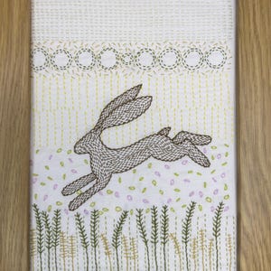 May include: A framed embroidery piece on white fabric featuring a brown stitched hare leaping through a field. The design includes green and yellow vertical stalks at the bottom, scattered pink and green leaf-like shapes, and a decorative circular border.