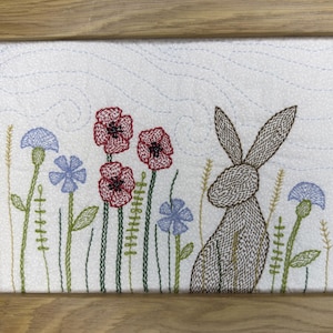 May include: A framed embroidery piece on white fabric featuring a brown stitched rabbit sitting among red poppies and blue cornflowers. The background includes light blue wavy stitched lines, and the entire artwork is enclosed in a light wood frame.