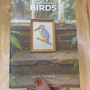 May include: A softcover book titled Kantha Inspirations Birds by Angela Daymond. The cover features a photograph of a framed embroidery of a blue and yellow kingfisher bird perched on a branch, set against a background of stone steps and plants.
