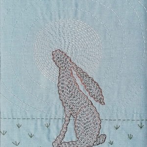 May include: An embroidered rabbit in brown, sitting on a blue background with green grass. The rabbit is looking up at a white circle with a radiating pattern.